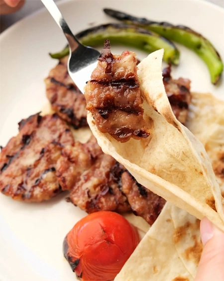 İzmir'in en beğenilen restoranlarından birinde sunulan et yemeklerinin iştah açıcı bir sunumu; ızgara izleri belirgin, sulu bir et parçası çatalda alınıp lavaş ekmeğine konulurken, tabağın geri kalanında közlenmiş domates ve biberlerle çevrili diğer ızgara köfteler lezzetli bir yemeği tamamlıyor.