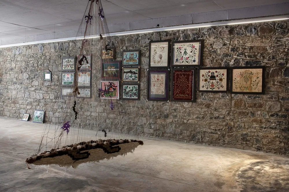 Baksı Sergi Salonu'nun rustik taş duvarları üzerinde çeşitli renklerde işleme ve dokuma sanat eserlerinin sergilendiği, tavanından sarkıtılmış büyük, ağ benzeri kahverengi ve bej tonlarında tekstil enstalasyonunun beton zeminin üzerinde süzüldüğü geniş bir sergi alanı manzarası.