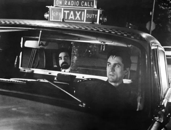 Martin Scorsese'nin yönettiği ikonik Taxi Driver filminden siyah beyaz bir karede, yönetmen Scorsese arka koltukta dikiz aynasında görünürken, Robert De Niro'nun canlandırdığı Travis Bickle karakteri taksi kullanırken düşünceli bir ifadeyle görülüyor.