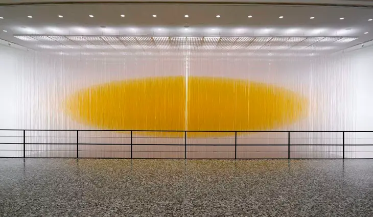Büyük ve modern bir galeri salonunda, Op Sanat akımının temelini oluşturan optik yanılsamaları barındıran, şeffaf dikey iplerden oluşan bir perdenin arkasında parlayan büyük, yatay elips şeklinde canlı sarı bir görüntü, adeta askıda süzülüyormuş gibi bir derinlik ve hareket hissi yaratıyor.