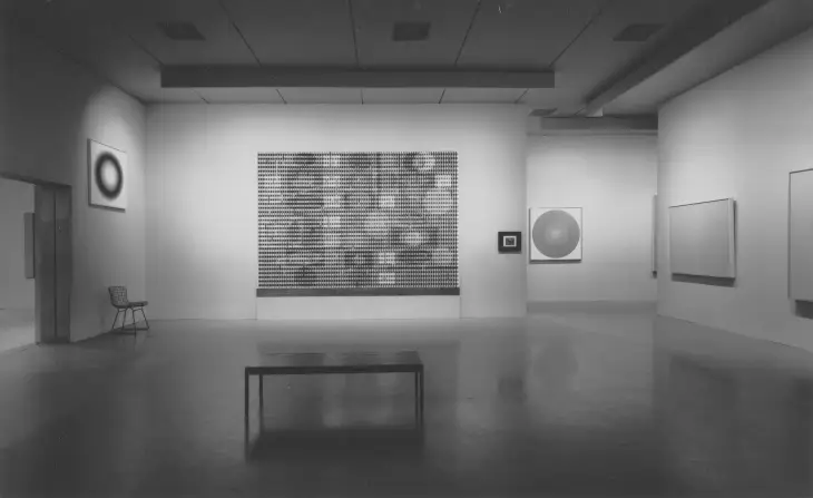 Gri tonlamalı modern bir sanat galerisi iç mekanında, Op Art sanat akımının karakteristik özelliklerini taşıyan, optik illüzyon yaratan büyük ızgara benzeri bir tablo merkezde yer alırken, çevresinde halka ve dairesel formlu diğer soyut eserler ile minimalist bir bank ve sandalye dikkat çekiyor.