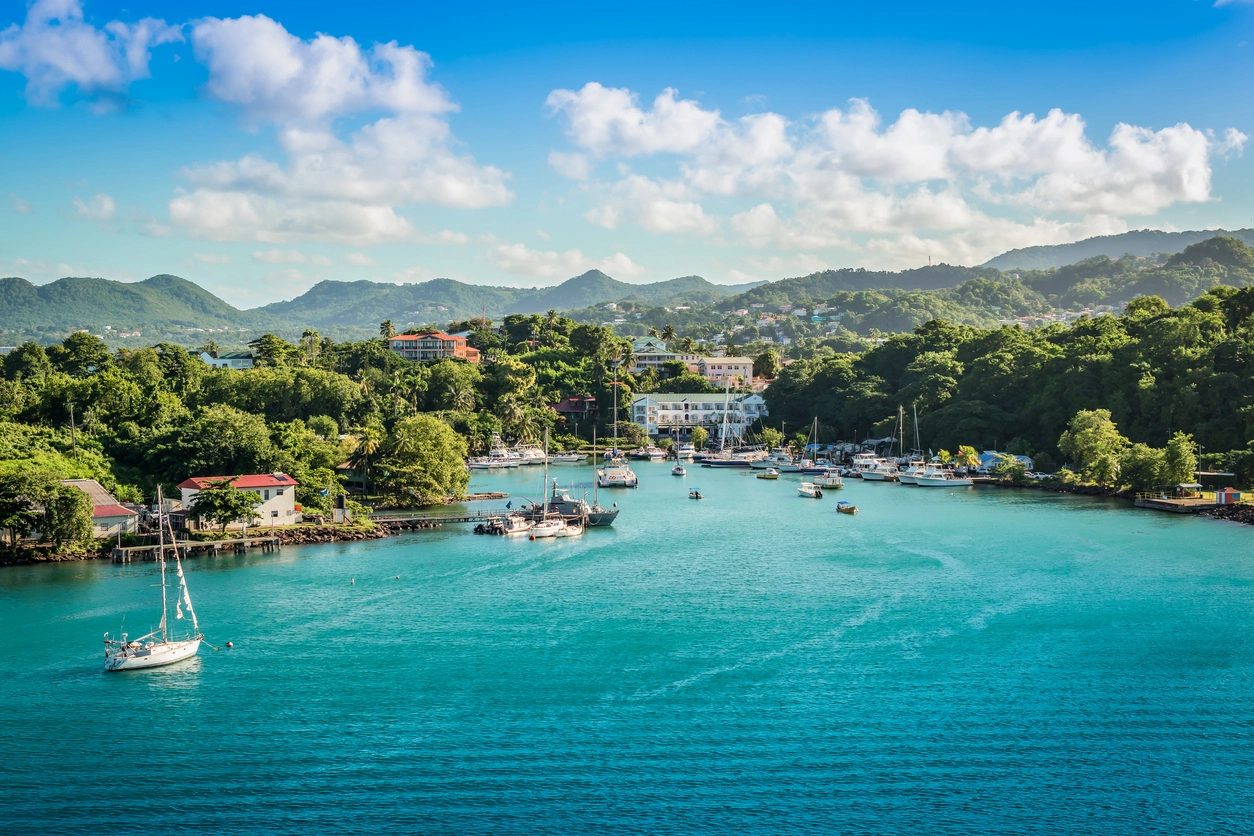 St. Lucia'nın ikonik Piton dağları ile çevrili, palmiye ağaçları ve yemyeşil bitki örtüsüyle dolu beyaz kumlu plajından turkuaz denize uzanan, ufukta küçük bir teknenin görüldüğü büyüleyici bir tropikal cennet manzarası.