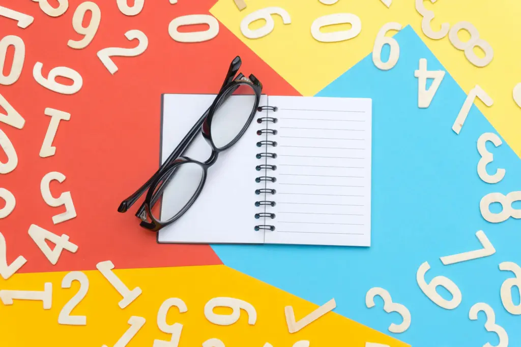 Renkli bir zeminde dağınık halde duran ahşap rakamlar, açık bir defter ve gözlükler... Sayıların büyülü dünyası numerolojiyi keşfetmeye hazır mısınız? Hayatınızın kodlarını çözmek için ilk adım.