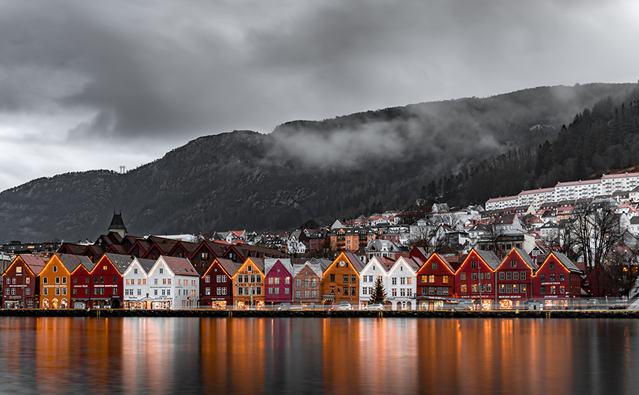 Norveç'in Bergen şehrindeki Bryggen limanında, karanlık dağlar ve bulutlu gökyüzü altında, ışıl ışıl parlayan rengarenk tarihi ahşap evlerin suya yansımasıyla oluşan masalsı manzara, Kışın Gidilecek En Güzel Yerler listesinden büyüleyici bir tatil rotası sunuyor.