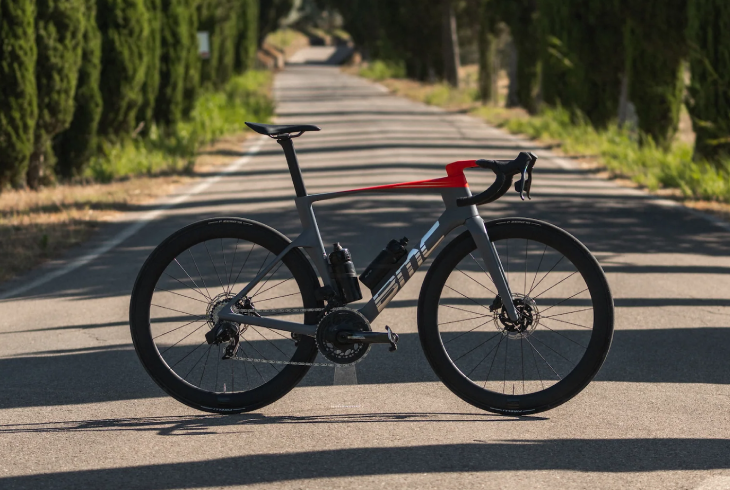 Ağaçların gölgelediği asfalt bir yolda duran, gri ve kırmızı renkleriyle dikkat çeken, disk frenli ve iki suluklu BMC Teammachine R yol bisikleti, aerodinamik performansı ve hafifliğiyle yarış odaklı sürüşler için en iyi bisiklet modellerinden biri olarak vurgulanıyor.