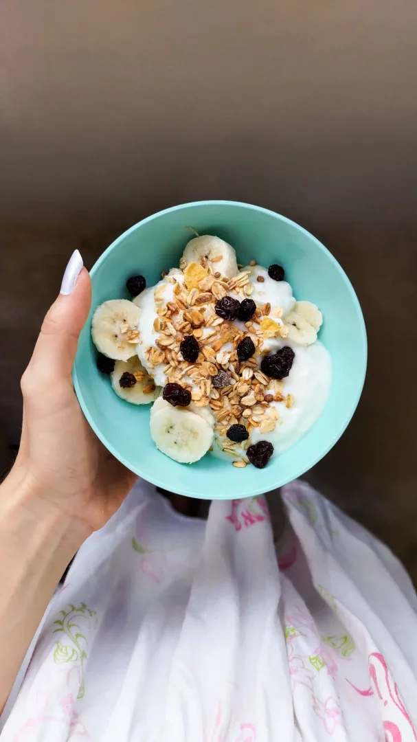 Beyaz ojeli bir elin tuttuğu, içinde dilimlenmiş muz, granola ve kuru üzüm ile süslenmiş açık mavi bir kase yoğurt, sağlıklı bir probiyotik kaynağı olarak fotoğrafın merkezinde yer alıyor.
