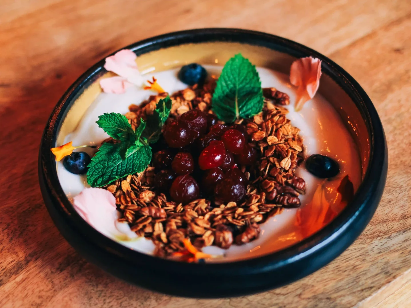 Koyu renkli bir kasenin içinde, probiyotik zengini, taze yoğurt üzerine serpiştirilmiş kıtır granola, koyu kırmızı taneli meyveler, parlak mavi yaban mersinleri, taze nane yaprakları ve renkli yenilebilir çiçek yapraklarıyla süslenmiş, sağlıklı ve göz alıcı bir tabak.