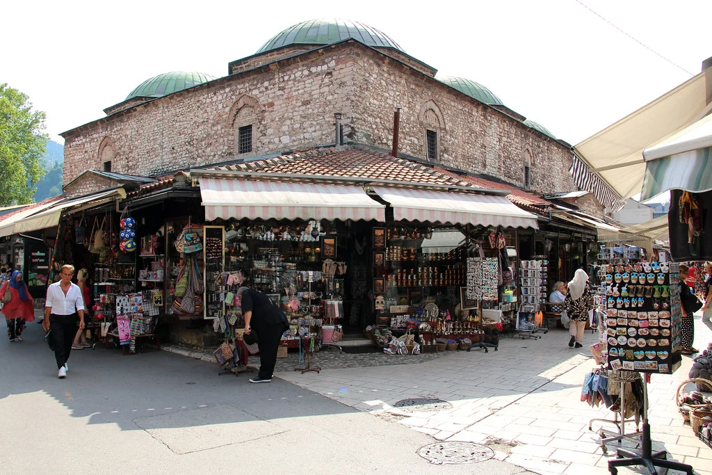 Yeşil renkteki kubbeleri ve taş duvarlarıyla tarihi bir yapının önünde uzanan, pembe beyaz çizgili tentelerin altındaki dükkanlarda çeşitli hediyelik eşyaların ve el sanatları ürünlerinin sergilendiği, kalabalık bir çarşıda insanlar alışveriş yaparken görülüyor.