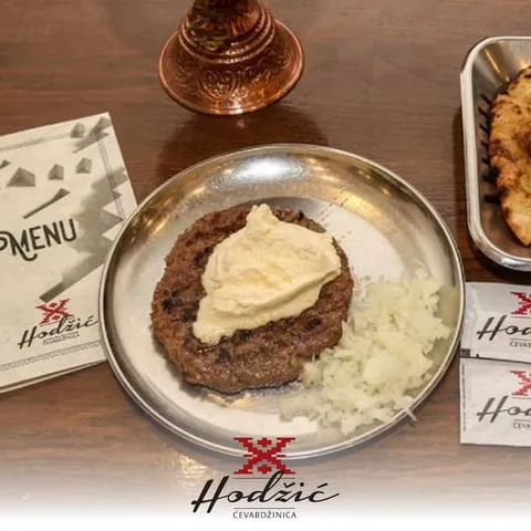 Bosna-Hersek gezi rehberinde Saraybosna'nın lezzet duraklarından biri olan Cevabdzinica Hodzic'te, üzerinde bolca kaymak ve yanında taze doğranmış soğanlarla servis edilen, metal bir tabakta duran geniş ızgara köfte, arka planda menü ve sıcak somun ekmeği ile geleneksel bir ziyafeti gözler önüne seriyor.
