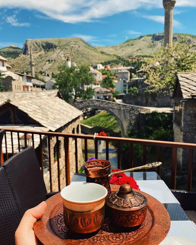 Mostar'ın tarihi dokusuyla iç içe, Kriva Ćuprija köprüsüne ve nehrin kıyısındaki geleneksel taş evlere nazır bir balkondan, bakır fincanında sunulan otantik Boşnak kahvesinin tadını çıkaran bir elin kadrajı, Bosna-Hersek gezisinin huzurlu anlarını yansıtıyor.