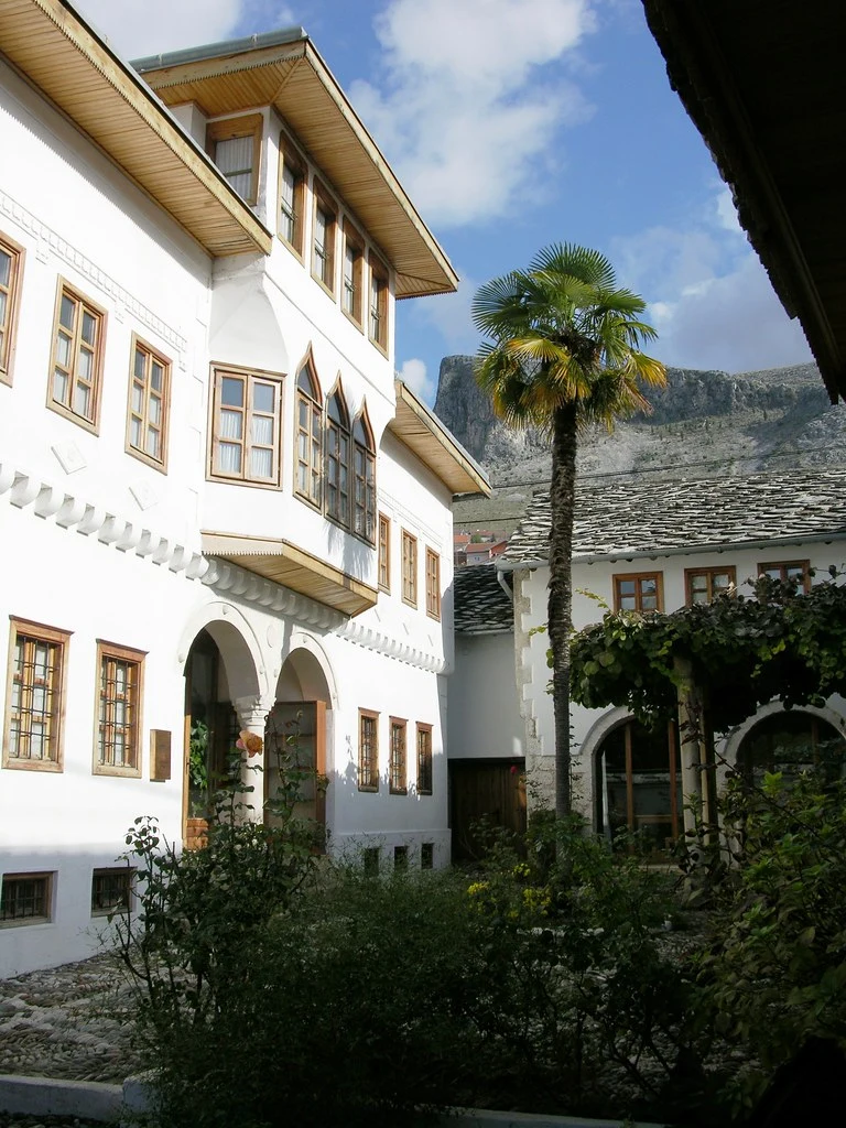 Mostar'ın tarihi Brankovac mahallesinde yer alan, geleneksel beyaz duvarlı Osmanlı konağı Muslibegović House'un ahşap saçaklı, kafesli pencereleri ve kemerli girişiyle bezeli dış avlusunu gösteren bu kare, Bosna-Hersek'in zengin kültürel mirasını doğal güzellikleriyle harmanlıyor; arkada yükselen dağ ve uzun palmiye ağacıyla huzurlu bir atmosfer sunuyor.