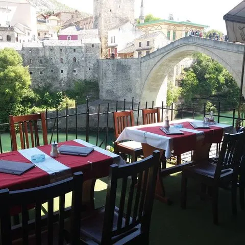 Kırmızı-beyaz örtülü masaları ve ahşap sandalyeleriyle şirin bir açık hava restoran terasından, Bosna-Hersek'in ikonik Mostar gezi rotasının önemli bir durağı olan yeşil Neretva Nehri üzerindeki tarihi Mostar Köprüsü ve çevresindeki eski taş yapılar net bir şekilde görülüyor.