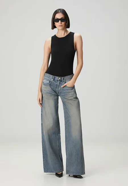 Siyah atlet ve güneş gözlüğü ile kombinlenen açık mavi, geniş paça wide leg jean pantolon; açık gri fon önünde modern ve iddialı bir stil sergiliyor.