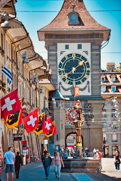 Bern’in ünlü Zytglogge Saat Kulesi ve İsviçre bayraklarıyla süslenmiş tarihi cadde – İsviçre gezi rehberi için ikonik manzara