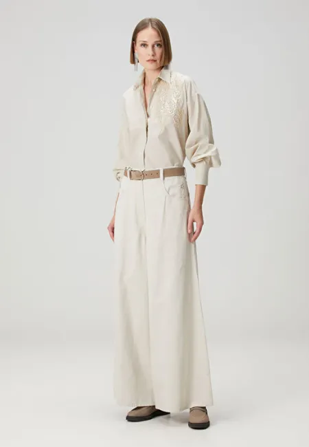 Ekru gömlek ve yüksek bel, geniş paça ekru wide leg jean pantolon giyen model; bej kemer ve düz ayakkabılarla açık gri fon önünde zarif ve minimal bir stil sergiliyor.