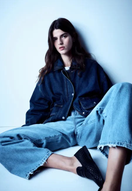 Koyu mavi oversize denim ceket ve açık mavi bol paça jean pantolon giyen model; siyah babetlerle mavi tonlu fon önünde cool ve rahat bir denim-on-denim stil sergiliyor.