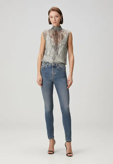 Yüksek yakalı, desenli kolsuz bluz ve mavi skinny jean pantolon giyen model; ince topuklu sandaletlerle açık gri fon önünde feminen ve zarif bir stil sergiliyor.