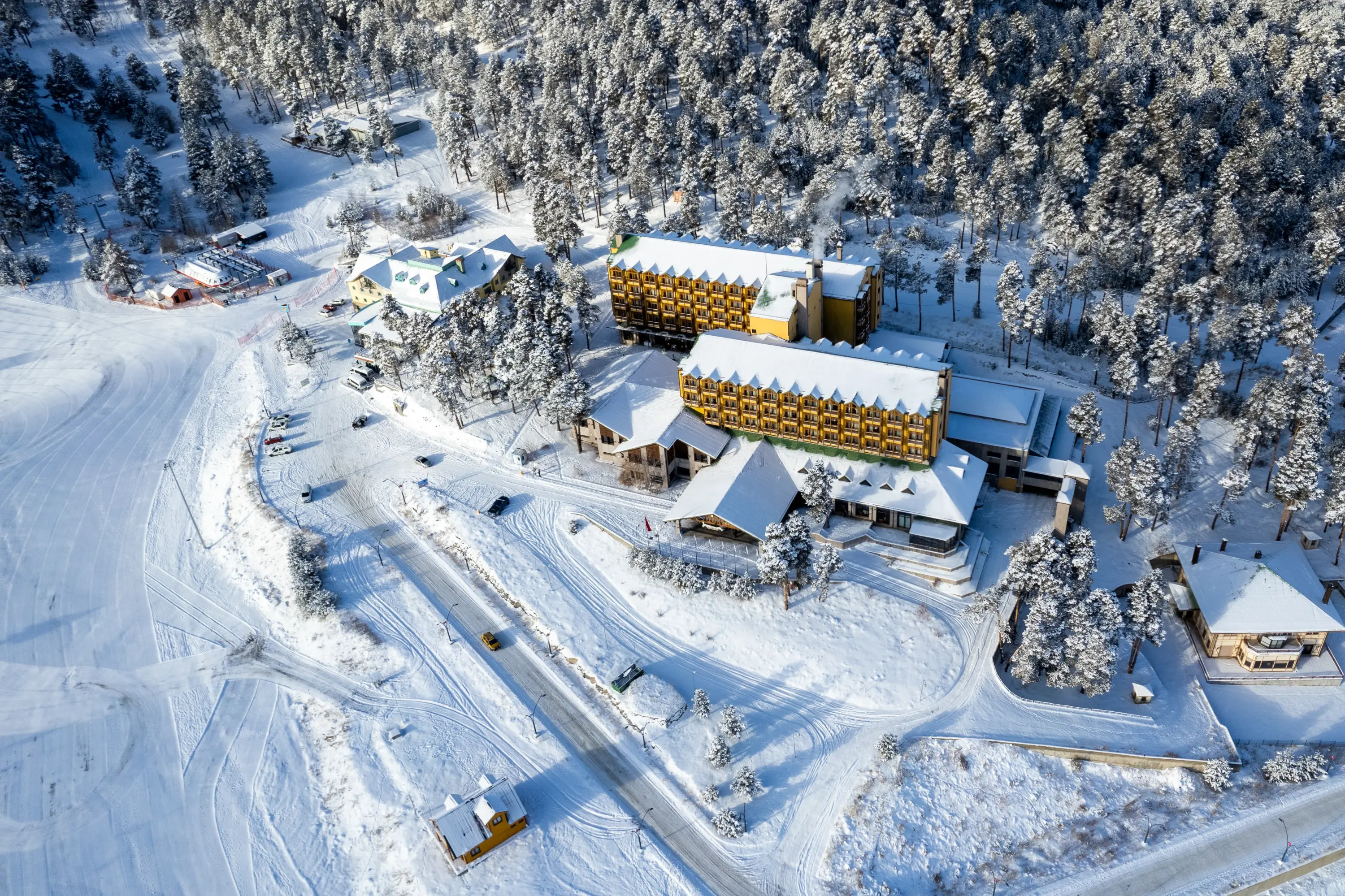 Kars Sarıkamış’ta çam ormanlarıyla çevrili kar manzarası içinde yer alan Duja Chalet Ski Resort’un havadan kış fotoğrafı; kayak pistine sıfır konumda, sarı renkli otel binası ve çevresindeki karla kaplı doğa görülüyor.