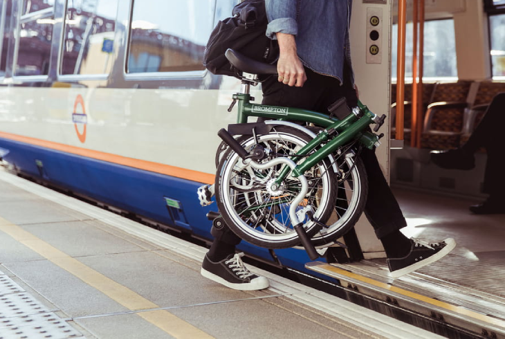 Yeşil bir Brompton katlanır bisikleti taşıyan bir kişi, şehir içi ulaşım ve toplu taşıma kolaylığını vurgulayarak bir tren platformunda trenin kapısından inerken veya binerken görülüyor, bu da katlanır bisikletlerin modern yaşamdaki pratikliğini gözler önüne seriyor.