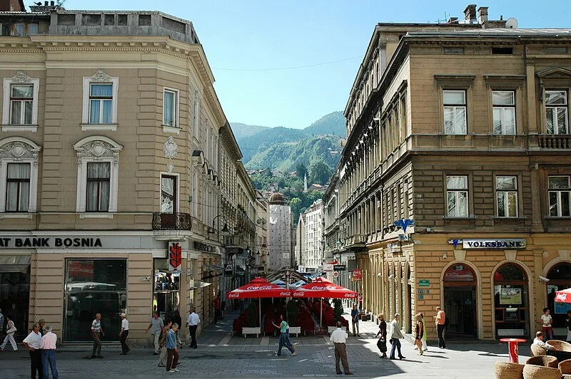 Sarajevo'nun tarihi Ferhadiye Caddesi'nde, eski mimariye sahip çok katlı binaların arasında uzanan, sağlı sollu bankaların ve ortada kırmızı şemsiyeli bir kafenin bulunduğu, birçok insanın yürüdüğü hareketli bir cadde manzarası, uzaklarda yemyeşil dağlarla çevrili şehrin kalabalık yaşamını ve doğal güzelliklerini bir araya getiriyor.
