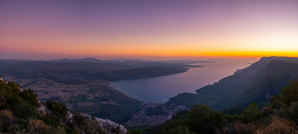 Gökova Körfezi'nin gün batımı veya doğumu anında çekilmiş panoramik manzarasında, mor ve turuncu tonlarındaki gökyüzü altında, sahil kasabası ve yemyeşil dağlar görülen bu etkileyici görüntü, bisiklet tutkunlarının mutlaka keşfetmesi gereken büyüleyici rotalardan birini gözler önüne seriyor.