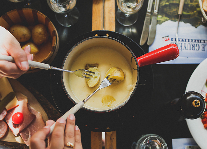 İsviçre seyahat rehberinin lezzet durağını yansıtan bu üstten çekilmiş karede, kırmızı saplı bir fondü tenceresinde erimiş peynire iki kişinin haşlanmış patates batırdığı an görülüyor; masada ayrıca şarküteri ürünleri ve ek patatesler ile ülkenin meşhur peynir fondü geleneği masalsı bir atmosferde sunuluyor.