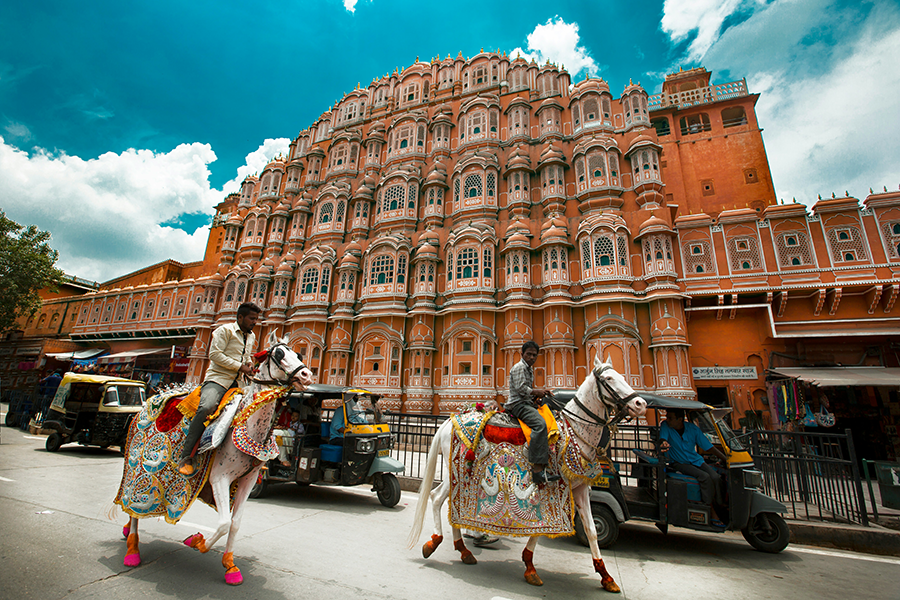 Hindistan'ın Jaipur şehrinde, süslü atlara binen iki adamın önünden geçtiği, pembe kumtaşından yapılmış, çok sayıda penceresi olan tarihi Hawa Mahal Sarayı, dünyanın en iyi bisiklet rotalarından biri olan Rajasthan bölgesinin kültürel zenginliğini yansıtan etkileyici bir mimari örneği.
