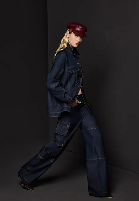 Koyu lacivert denim ceket ve yüksek bel jean kargo pantolon giyen model; bordo şapka ve topuklu ayakkabılarla koyu arka plan önünde güçlü ve maskülen bir stil sergiliyor.