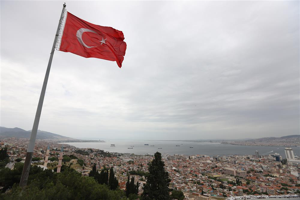 Yüksek bir noktadan çekilmiş bu geniş açılı görselde, bulutlu bir gökyüzü altında uzun bir direkte dalgalanan Türk bayrağı, ön planda şehri, körfezde yüzen gemileri ve uzaklardaki dağları kapsayan İzmir'in etkileyici panoramasını gözler önüne seriyor.