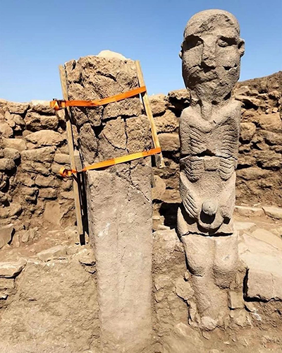 Karahantepe'nin arkeolojik kazı alanında, belirgin kaburgaları ve ellerini karnında kavuşturmuş şekilde duran, insanlık tarihine ışık tutan antik bir insan heykeli, hemen yanında ahşap desteklerle koruma altına alınmış dikdörtgen bir dikilitaş ile birlikte, bu gizemli yerleşim yerinin önemli buluntularını gözler önüne seriyor.