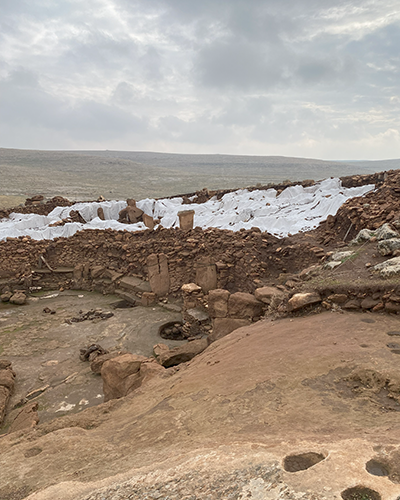 Tarihin yeni gizemli noktası Karahantepe'deki kazı alanının geniş bir görünümünde, beyaz örtülerle korunan Neolitik döneme ait yapılar ve dikilitaşlar, taşlık, kahverengi zeminde dairesel oyuklar ve bulutlu gökyüzü altında uzanan boş arazi görülüyor.