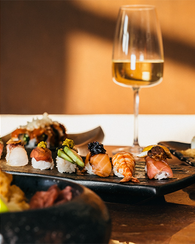 Yakın plandan çekilen bu karede, siyah bir servis tabağında özenle dizilmiş somon, karides ve avokado gibi farklı malzemelerle hazırlanan rengarenk nigiri ve maki sushiler ile arka planda, ortamın şıklığını vurgulayan bir kadeh beyaz şarap, İzmir'in en beğenilen sushi restoranlarından birindeki üst düzey bir lezzet deneyimini gözler önüne seriyor.