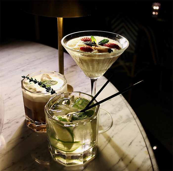 Loş ışıklı bir barda veya restoranda mermer bir yüzeyde sergilenen üç farklı içecek: çilek ve nane yaprağı süslemeli kremalı bir martini kadehi, limon dilimleri, nane yaprakları ve buzla dolu şeffaf, köşeli bir kokteyl bardağı ile üzeri krem şanti ve taze kekik dallarıyla süslü, kahvemsi tonda bir başka kokteyl, arka plandaki altın renkli masa lambasının sıcak ışığı altında parlıyor.