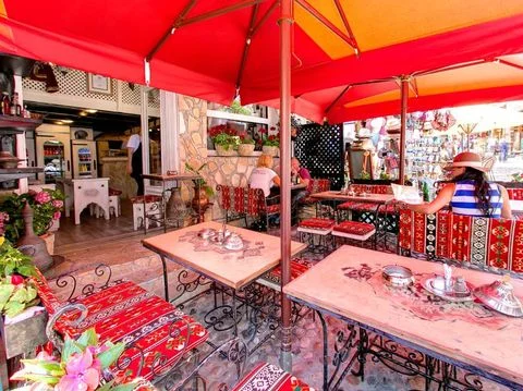Geleneksel Bosna motifli kırmızı örtülerle döşenmiş sandalyeler ve masalarıyla, Mostar'ın Eski Şehir'indeki Tima-Irma Restoranı'nın hareketli terasında, kırmızı ve turuncu şemsiyelerin altında oturan misafirler, şehrin canlı atmosferini deneyimliyor.