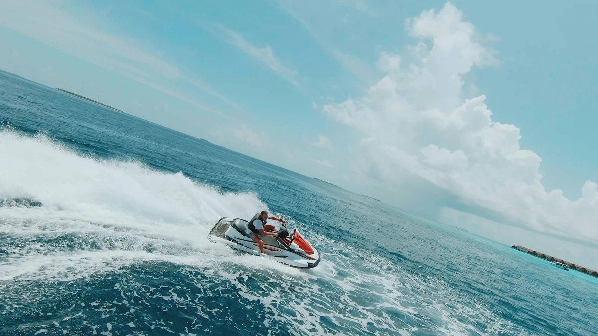 Parlak güneşli bir günde, masmavi denizde beyaz ve turuncu renkli bir jet ski ile hızla ilerleyen can yelekli bir kişinin arkasında büyük bir köpüklü dalga oluşturduğu dinamik bir an, su sporları için doğru ekipman seçiminin önemini vurguluyor.