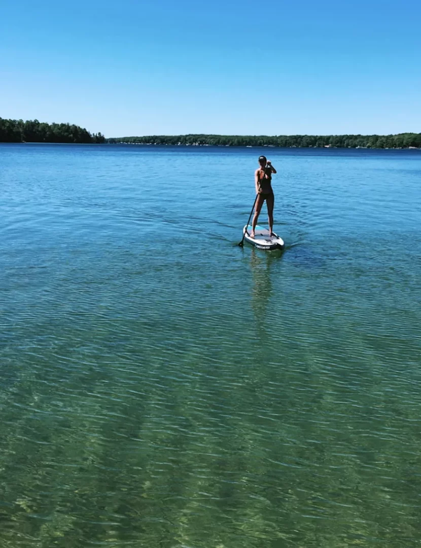 Berrak, turkuaz sular üzerinde dengede duran, siyah mayolu bir kadın, Stand Up Paddle (SUP) tahtasıyla dingin bir gölde kürek çekerek ilerlerken, arkasında yemyeşil ormanlarla kaplı uzak bir kıyı şeridi ve masmavi bir gökyüzü dikkat çekiyor.