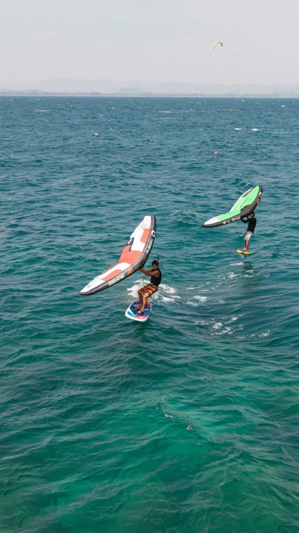 Mavi-yeşil dalgalı sular üzerinde iki sporcu wing foil yapıyor; öndeki sporcu kırmızı-beyaz bir kanatla suya yakın ilerlerken, arkadaki sporcu yeşil kanatla suyun üzerinde yükseliyor, bu dinamik sahne Türkiye'de wing foil sporunun heyecanını yansıtıyor.