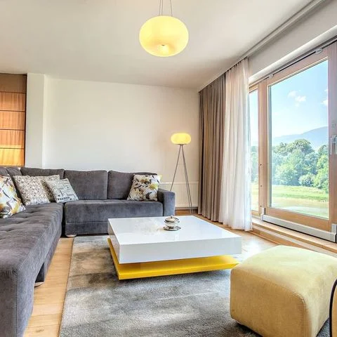 Saraybosna'daki bir VIP Apartments dairesinin, konforlu L-şeklinde gri kanepesi, sarı detaylı modern sehpası ve yerden tavana uzanan penceresinden görünen yemyeşil Bosna-Hersek doğa ile çevrili dağ manzarasıyla hem ev sıcaklığını hem de lüks bir konaklama deneyimini bir araya getiren ferah ve şık bir oturma odası.