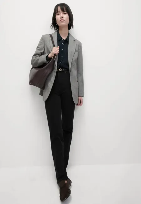 Gri blazer ceket, koyu gömlek ve yüksek bel siyah straight leg jean pantolon kombinli model; omzunda büyük kahverengi çanta ile açık gri fon önünde şık ve maskülen bir stil sergiliyor.