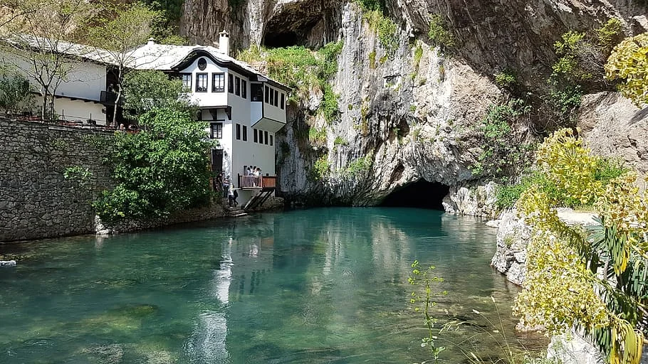 Bosna-Hersek'in tarihi Blagaj Tekkesi'nin yer aldığı bu manzarada, kayalık bir yamaca yaslanmış geleneksel beyaz yapı, turkuaz renkli berrak sularıyla dikkat çeken bir nehrin kenarında duruyor, nehir ise sağdaki büyük bir mağara girişinden fışkırarak etkileyici bir doğal güzellik sunuyor.