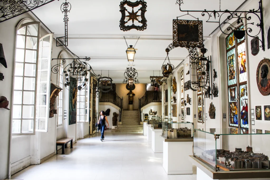 Parlak beyaz duvarlar ve zemine sahip geniş bir müze galerisinde, tavandan ve duvarlardan sarkan işlemeli ferforje tabelalar, duvarları süsleyen vitray pencereler ve çeşitli sanatsal eserler, ziyaretçilere Paris tarihini yansıtan zengin bir koleksiyon sunarken, ilerideki merdivenlere doğru yürüyen bir kişi ve zeminde sergilenen mimari modeller dikkat çekiyor.