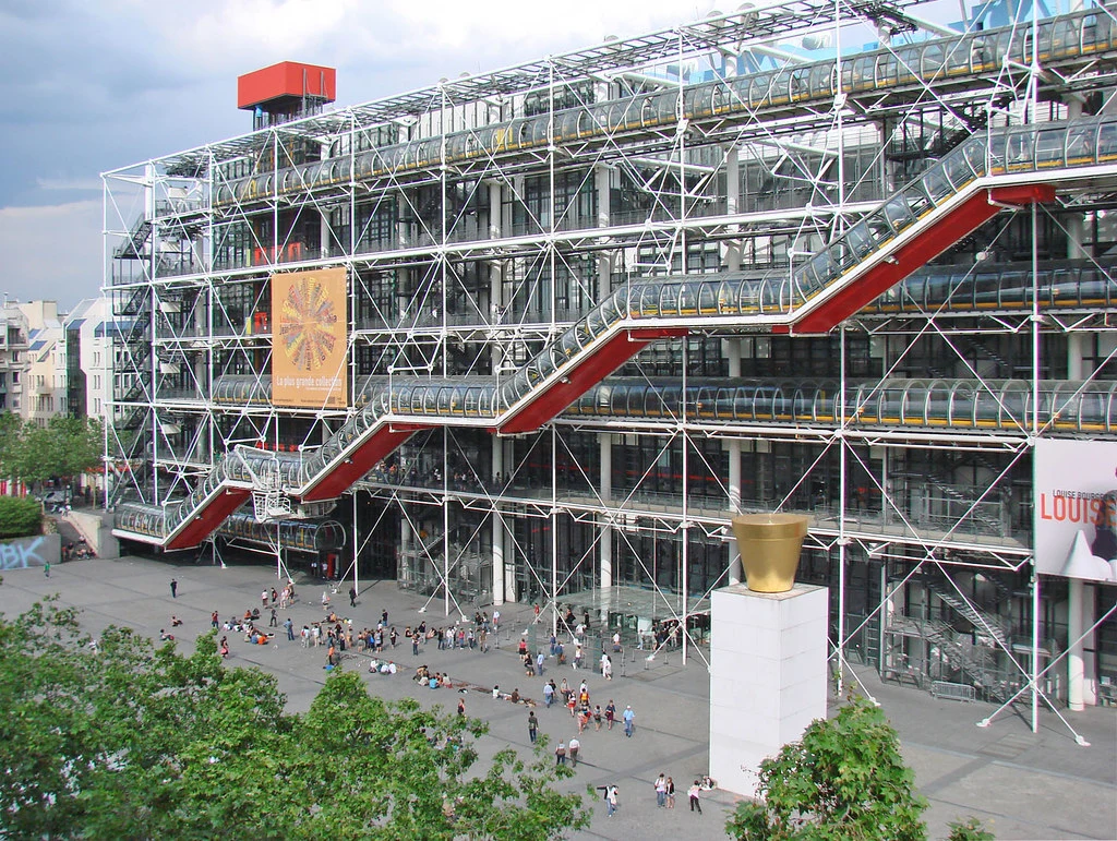 Paris'teki ikonik Centre Pompidou binası, dış cephesindeki açıkta duran renkli boruları, kırmızı cam tüpler içindeki yürüyen merdivenleri ve yapısal elemanlarıyla, önündeki hareketli meydanda toplanmış insanlarla ve beyaz bir kaide üzerindeki büyük altın renkli bir heykelle birlikte, bulutlu bir günde geniş bir açıyla sergileniyor.