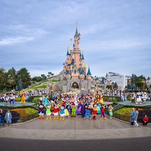 Pembe ve mavi kulelere sahip Disneyland Paris Şatosu'nun önünde, Mickey Mouse, Donald Duck, prensesler ve çeşitli Disney karakterlerinden oluşan coşkulu bir kalabalık, ellerini havaya kaldırarak düzenlenmiş bahçelerle çevrili geniş bir alanda neşeyle poz veriyor.