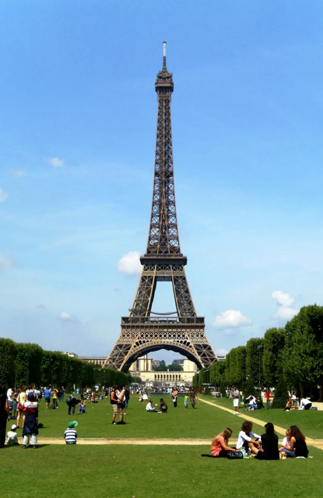 Eyfel Kulesi'nin heybetli siluetinin, güneşli bir Paris gününde yemyeşil Champ de Mars çimlerinde oturan ve yürüyen kalabalıklar arasında yükseldiği, her köşesi bir film sahnesi Paris gezi rehberinin ikonik duraklarından birini gösteren canlı bir manzara.