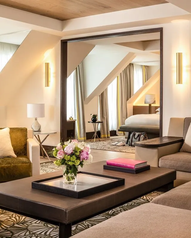 Paris gezi rehberi kapsamında sunulan lüks konaklama seçeneklerinden birini yansıtan bu şık otel süiti, eğimli tavanları, duvar apliklerinden yayılan sıcak ışıkları, üzerinde aynalı tepside canlı bir çiçek buketi ve parlak pembe bir kitabın yer aldığı geniş sehpasıyla davetkar bir atmosfer sunuyor.
