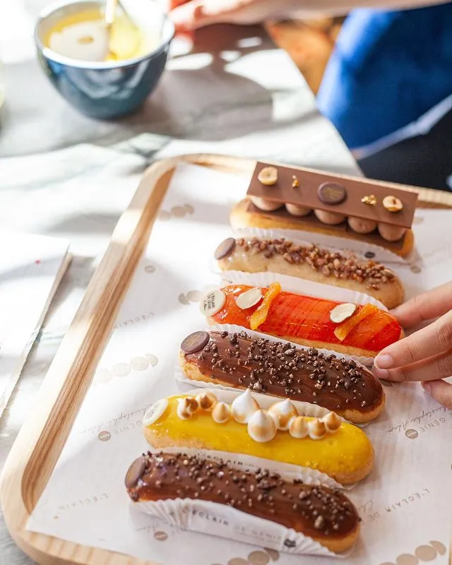 Bir Paris gezi rehberinin öne çıkardığı L'Éclair de Génie pastanesinden ahşap bir tepside sunulan, karamel, çikolata, parlak turuncu narenciye ve canlı sarı limon kreması gibi farklı dolgularla modern dokunuşlarla süslenmiş çeşitli eklerler, Paris'in zengin pastane kültürünü yansıtıyor.