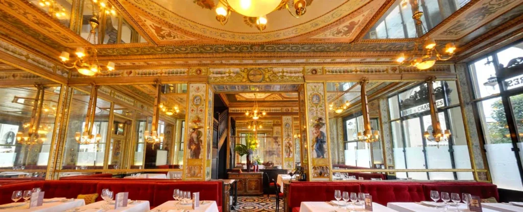 Le Grand Véfour restoranının göz alıcı iç mekanında, altın yaldızlı detaylarla süslenmiş işlemeli tavanlar, zarif avizeler, odanın genişliğini yansıtan aynalı duvarlar ve rahat kırmızı kadife oturma gruplarıyla donatılmış beyaz masa örtülü masalar yer alıyor.
