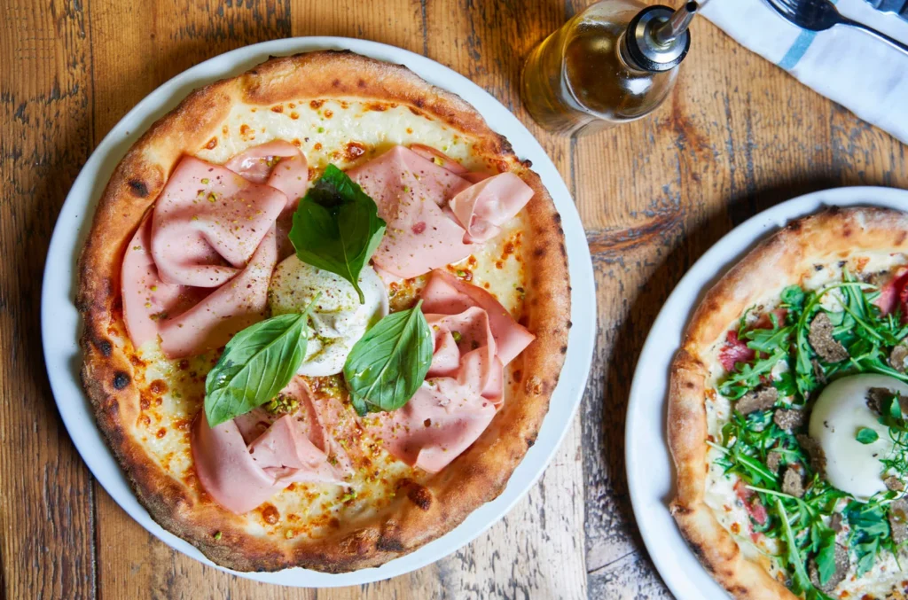 Little Nonna'dan servis edilen, üzeri pembe mortadella dilimleri, taze burrata peyniri, yeşil fesleğen ve fıstık kırıntılarıyla süslenmiş, kenarları hafif yanık ve çıtır bir İtalyan pizzası ile yanında trüf mantarlı başka bir pizza, Paris gezi rehberinde öne çıkan glutensiz İtalyan mutfağı deneyimini yansıtıyor.