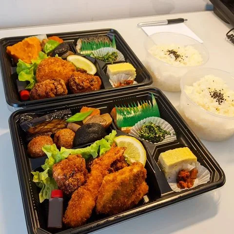 Siyah renkli iki adet bento kutusu, her biri kızarmış karides, tavuk parçaları, yeşil salata, limon dilimi, tamagoyaki ve çeşitli sebzelerle dolu; yanında iki şeffaf kapta susamlı pilav bulunan, hazır bir Japon yemeği setini gösteriyor.
