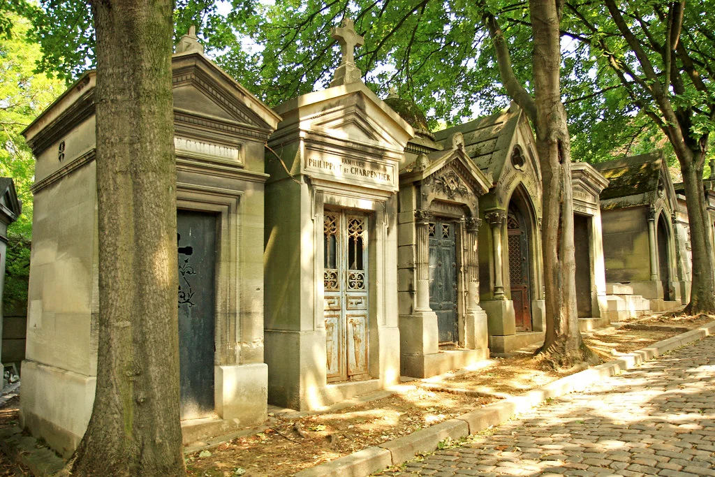 Père Lachaise Mezarlığı'ndan bir görünüm: Yeşil yapraklı ağaçların gölgesinde, parke taşlı bir yolun kenarında uzanan, süslü kapıları ve işlemeli taş yapılarıyla eski ve görkemli mozolelerden oluşan uzun bir sıra görülüyor, hafif güneş ışığı yolu aydınlatıyor.